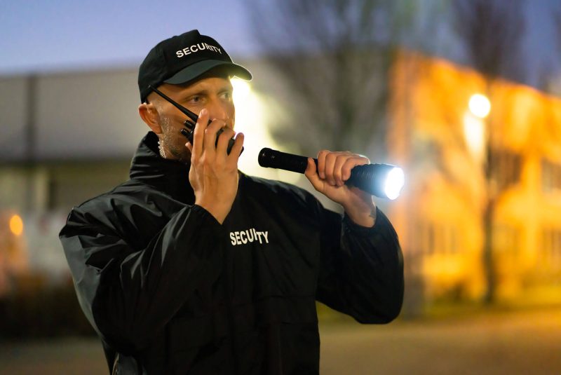 Uomo in uniforme di sicurezza con radio e torcia in un'area esterna illuminata.