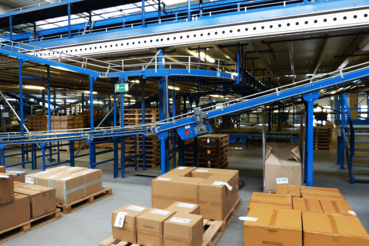 Interno di magazzino con nastri trasportatori blu, pacchi e pallet impilati.